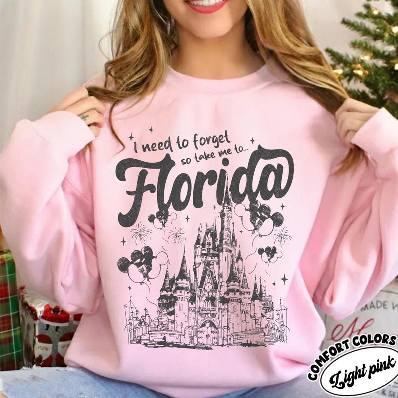 May include: Pale pink sweatshirt featuring the words "I need to forget so take me to... Florida" in black. The design includes a castle, fireworks, and Mickey Mouse balloons. The sweatshirt is labelled "Comfort Colors".
