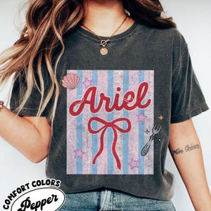 May include: Dark gray Comfort Colors t-shirt featuring a graphic of Ariel with a red bow, seashell, and stars on a pink and blue striped background. The word "Ariel" is in red script.