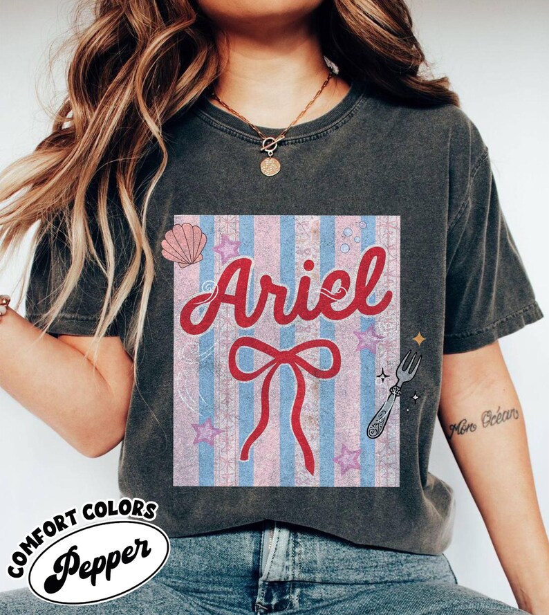May include: Dark gray Comfort Colors t-shirt featuring a graphic of Ariel with a red bow, seashell, and stars on a pink and blue striped background. The word "Ariel" is in red script.