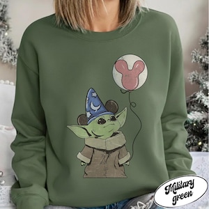 May include: Military green sweatshirt featuring a cartoon character wearing a wizard hat and holding a Mickey Mouse balloon. The character is green with large ears and a brown coat. The balloon is pink and white. The text "Military green" is in a white oval.