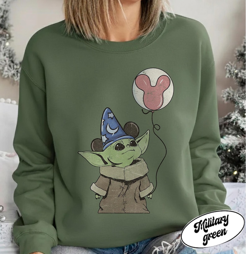 May include: Military green sweatshirt featuring a cartoon character wearing a wizard hat and holding a Mickey Mouse balloon. The character is green with large ears and a brown coat. The balloon is pink and white. The text "Military green" is in a white oval.