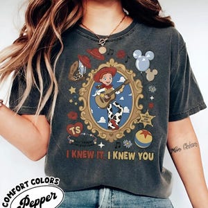 May include: A charcoal gray short-sleeve t-shirt featuring a central graphic of a cowgirl playing a guitar inside an ornate gold frame. Surrounding elements include a cowboy hat, a boot, balloons, a star badge, and the text I KNEW IT, I KNEW YOU.