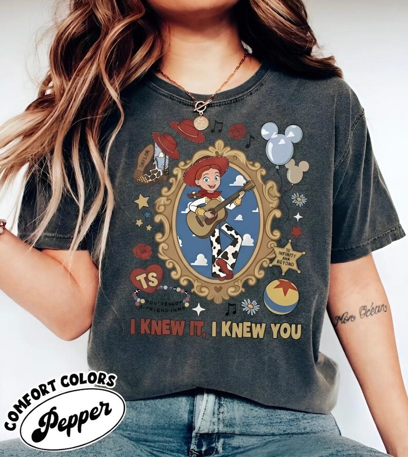 May include: A charcoal gray short-sleeve t-shirt featuring a central graphic of a cowgirl playing a guitar inside an ornate gold frame. Surrounding elements include a cowboy hat, a boot, balloons, a star badge, and the text I KNEW IT, I KNEW YOU.