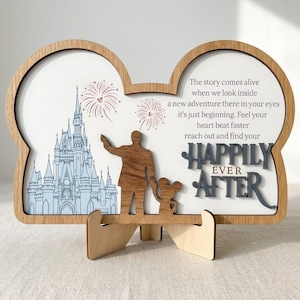 May include: A wooden plaque shaped like mouse ears featuring a light blue castle illustration, two firework designs, and a silhouette of a person holding a child's hand. The text reads: The story comes alive when we look inside a new adventure there in your eyes it’s just beginning. Feel your heart beat faster reach out and find your HAPPILY EVER AFTER.