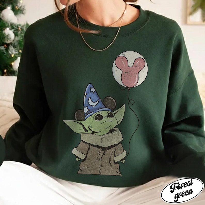 May include: Forest green sweatshirt featuring a cartoon character with large ears, wearing a wizard hat and a brown coat. The character is holding a balloon with a Mickey Mouse-shaped design. The words "Forest green" are in a white oval.