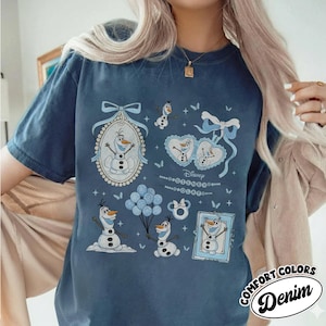 Pode incluir: Camiseta azul denim com ilustrações de Olaf de Frozen da Disney. O design inclui imagens emolduradas, corações, balões e borboletas. A camisa tem as palavras "Comfort Colors" e "Denim" impressas.