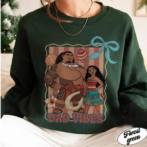 May include: Forest green sweatshirt featuring a graphic of two cartoon characters with the text "DAD VIBES". The graphic is framed with a light brown border and includes a blue bow and tropical elements.