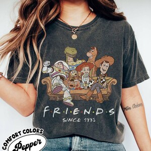 May include: Dark gray t-shirt featuring a graphic of Toy Story characters sitting on a couch. The word "FRIENDS" is printed below the graphic, with "SINCE 1995" underneath. The shirt is from Comfort Colors.