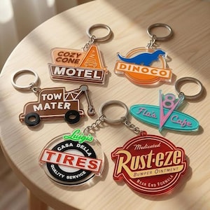Puede incluir: Seis llaveros de acr&iacute;lico con dise&ntilde;os coloridos inspirados en temas automotrices. Los llaveros muestran textos que incluyen Cozy Cone Motel, Dinoco, Tow Mater, Flo's V8 Cafe, Luigi's Casa Della Tires y Rust-eze Bumper Ointment con anillas.