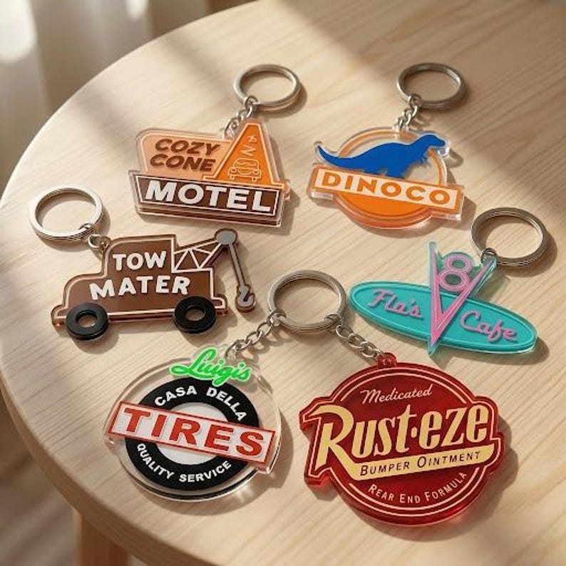 Puede incluir: Seis llaveros de acr&iacute;lico con dise&ntilde;os coloridos inspirados en temas automotrices. Los llaveros muestran textos que incluyen Cozy Cone Motel, Dinoco, Tow Mater, Flo's V8 Cafe, Luigi's Casa Della Tires y Rust-eze Bumper Ointment con anillas.