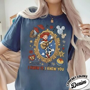 May include: A blue short-sleeve t-shirt featuring a graphic of a cowgirl playing a guitar inside an ornate gold frame. The design includes a cowboy hat, balloons, a star badge, a ball, and the text "I KNEW IT, I KNEW YOU" in yellow letters.