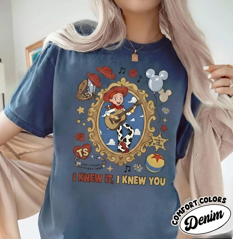 May include: A blue short-sleeve t-shirt featuring a graphic of a cowgirl playing a guitar inside an ornate gold frame. The design includes a cowboy hat, balloons, a star badge, a ball, and the text "I KNEW IT, I KNEW YOU" in yellow letters.