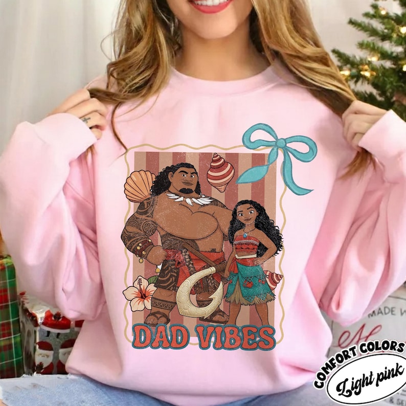 May include: Light pink sweatshirt featuring a graphic of two cartoon characters with the text "DAD VIBES". The design includes a bow, seashells, and a flower. The sweatshirt is labeled "COMFORT COLORS" and "Light pink".