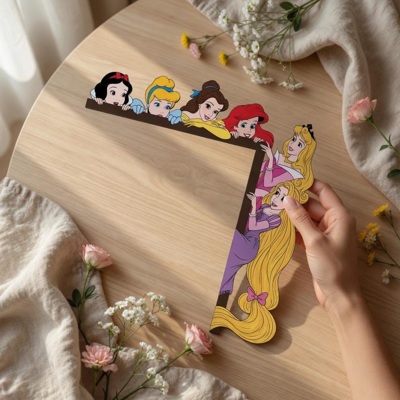May include: A decorative corner frame featuring a series of illustrated cartoon princesses including Snow White, Cinderella, Belle, Ariel, Aurora, and Rapunzel. The characters are arranged along the top and right edges of the L-shaped wooden border.