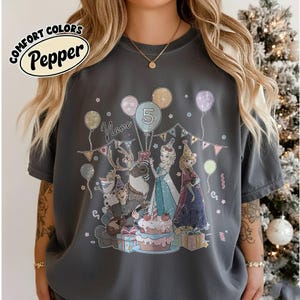 Puede incluir: Una camiseta Comfort Colors gris oscuro con un gr&aacute;fico de cumplea&ntilde;os colorido. El gr&aacute;fico presenta personajes de dibujos animados, globos, un pastel y el n&uacute;mero 5. La camiseta tambi&eacute;n tiene la palabra "Name" encima del gr&aacute;fico.