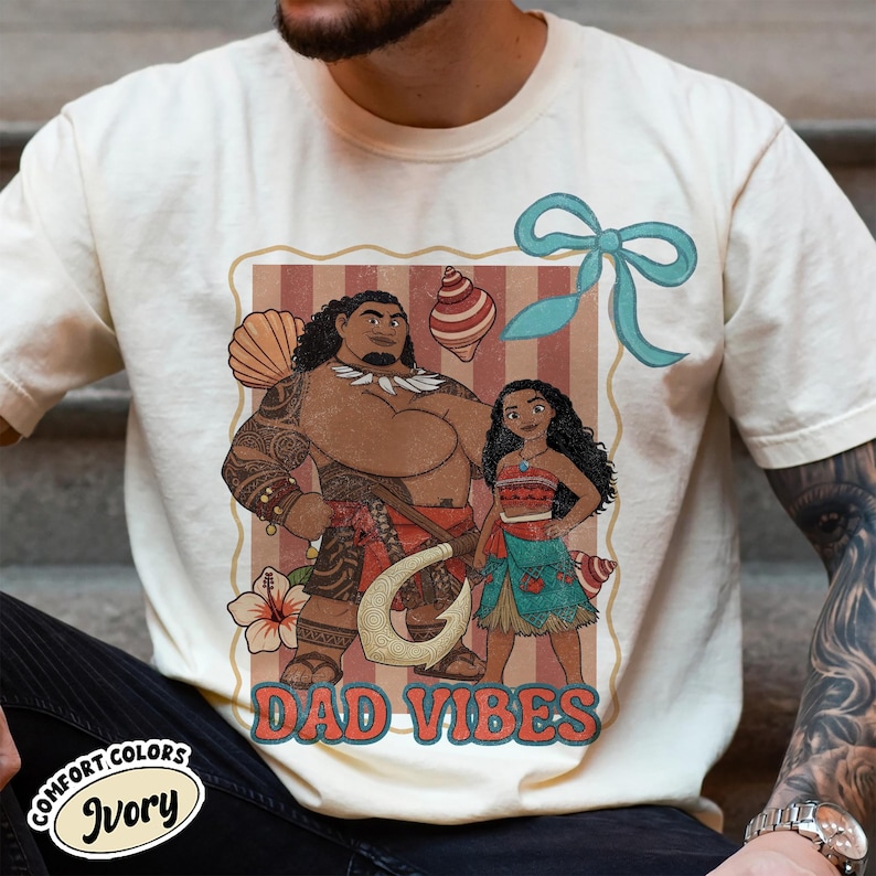 May include: Ivory-colored t-shirt featuring a cartoon illustration of two characters with the text "DAD VIBES". The graphic includes a man with tattoos and a woman, along with seashells and a blue bow. The shirt is labeled "Comfort Colors Ivory".
