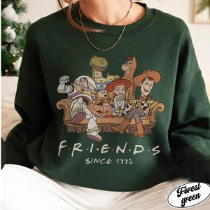 May include: Forest green sweatshirt featuring a graphic of Toy Story characters on a couch. The word "FRIENDS" is printed in colorful letters, with "SINCE 1995" below. The sweatshirt is a solid color with a crew neck.