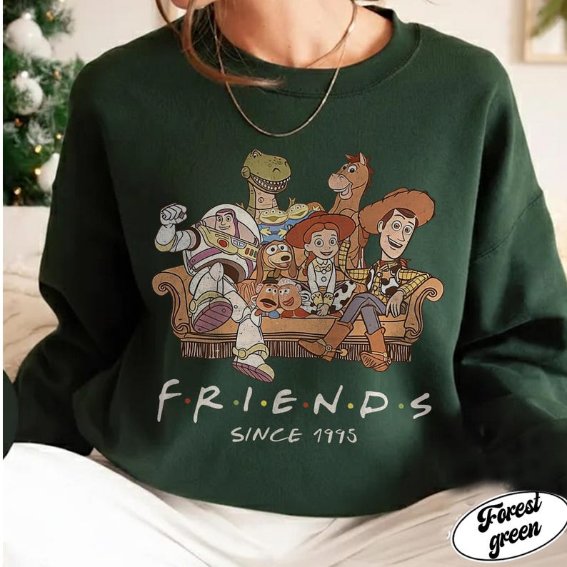 May include: Forest green sweatshirt featuring a graphic of Toy Story characters on a couch. The word "FRIENDS" is printed in colorful letters, with "SINCE 1995" below. The sweatshirt is a solid color with a crew neck.
