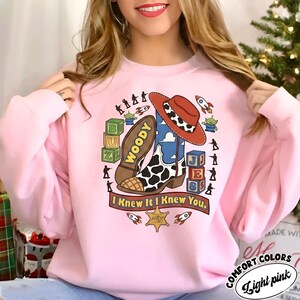 May include: A light pink crewneck sweatshirt featuring a graphic print of a cowboy boot, a cow-print hat, alphabet blocks spelling BUZ and JES, green alien figures, toy soldiers, rockets, and a sheriff badge with the text I Knew It I Knew You.
