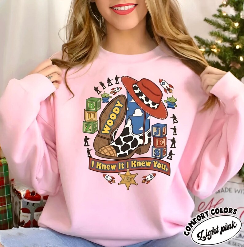 May include: A light pink crewneck sweatshirt featuring a graphic print of a cowboy boot, a cow-print hat, alphabet blocks spelling BUZ and JES, green alien figures, toy soldiers, rockets, and a sheriff badge with the text I Knew It I Knew You.