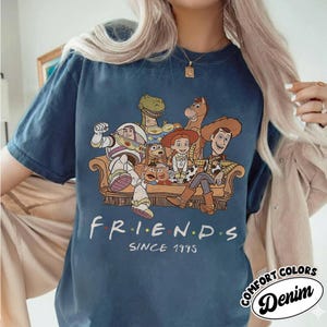 May include: A navy blue t-shirt featuring a graphic of Toy Story characters sitting on a couch. The word "FRIENDS" is printed below the characters, with "SINCE 1995" underneath. The shirt also has the words "COMFORT COLORS Denim" printed on it.