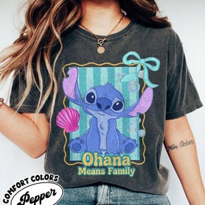 May include: Dark gray Comfort Colors t-shirt featuring a blue cartoon character with large ears and the text "Ohana Means Family." The design includes a pink seashell and a light blue bow. The shirt has a vintage, worn look.