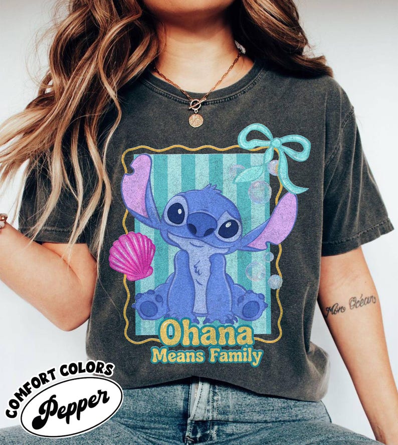 May include: Dark gray Comfort Colors t-shirt featuring a blue cartoon character with large ears and the text "Ohana Means Family." The design includes a pink seashell and a light blue bow. The shirt has a vintage, worn look.