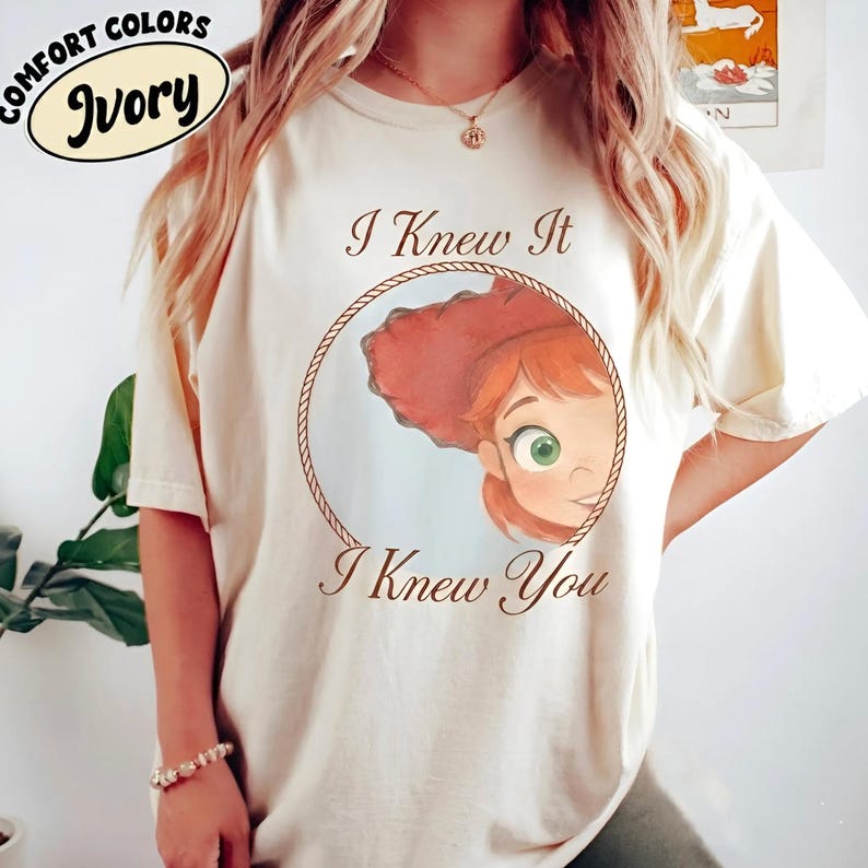 May include: An ivory short-sleeve t-shirt featuring a graphic of a cartoon character with red hair and green eyes inside a rope-style circle. The text "I Knew It" appears above the circle and "I Knew You" appears below in brown script lettering.