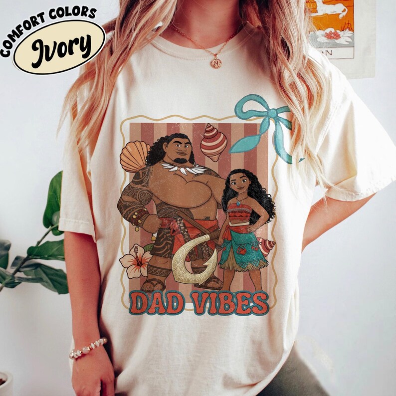 May include: Ivory-colored t-shirt with a graphic featuring two cartoon characters and the words "DAD VIBES." The graphic includes a brown-skinned man with tattoos and a woman in a blue and red outfit. The shirt also has the words "COMFORT COLORS" and "Ivory."