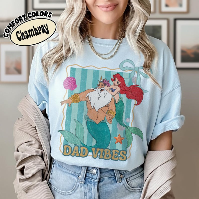 May include: Light blue t-shirt with a graphic of a mermaid and a merman with the text "DAD VIBES". The shirt also has the text "COMFORT COLORS Chambray" in a circular design. The graphic is in a rectangular frame with blue stripes.