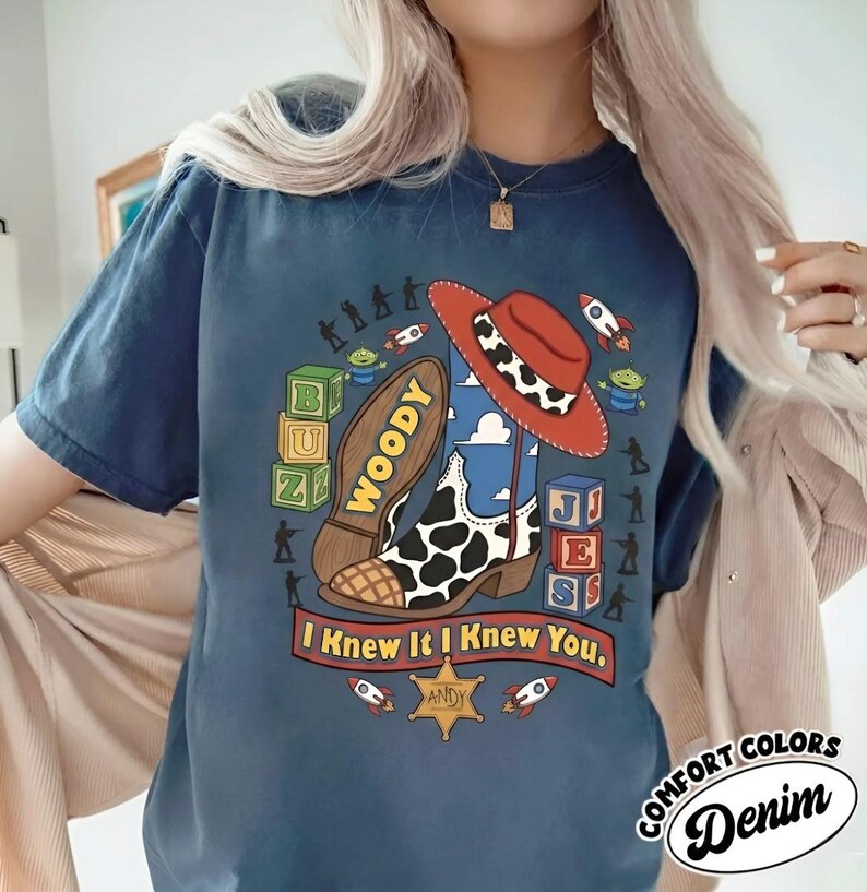 May include: A dark blue t-shirt featuring a graphic print of a cowboy boot, a red cowboy hat, alphabet blocks spelling BUZ and JES, small green aliens, rockets, and a sheriff badge with the text ANDY. The text I Knew It I Knew You is printed below.