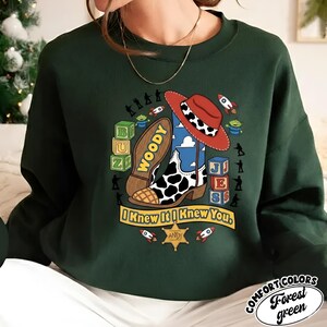 May include: A forest green crewneck sweatshirt featuring a graphic print of a cowboy boot with a red hat, toy blocks, rockets, and small soldier silhouettes. The text reads "WOODY" on the boot sole, "I Knew It I Knew You" on a banner, and "ANDY" on a star badge.