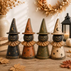 May include: Four decorative wooden witch figurines standing in a row. Each figure features a pointed hat, a round pumpkin-like head with a carved face, and a hessian ruffle collar with a small crescent moon charm. They are painted in black, orange, olive green, and cream, decorated with stars and moon motifs.