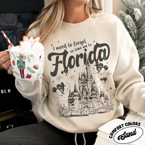 May include: Sand-coloured sweatshirt featuring the text "I need to forget so take me to Florida" and a graphic of a castle, fireworks, and Mickey Mouse balloons. Made from Comfort Colors material.
