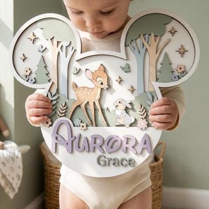 May include: A layered wooden wall sign shaped like mouse ears featuring a fawn and a rabbit in a forest setting. The sign includes the name Aurora in large purple script and the name Grace in smaller grey text, accented with stars and flowers.