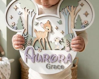 May include: A layered wooden wall sign shaped like mouse ears featuring a fawn and a rabbit in a forest setting. The sign includes the name Aurora in large purple script and the name Grace in smaller grey text, accented with stars and flowers.