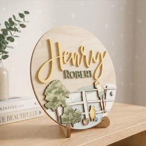 May include: A circular wooden sign featuring the name Henry Robert in raised yellow and green lettering. The design includes a layered wooden scene of a person and a bear leaning on a white fence near green trees, all set on a light wood base.