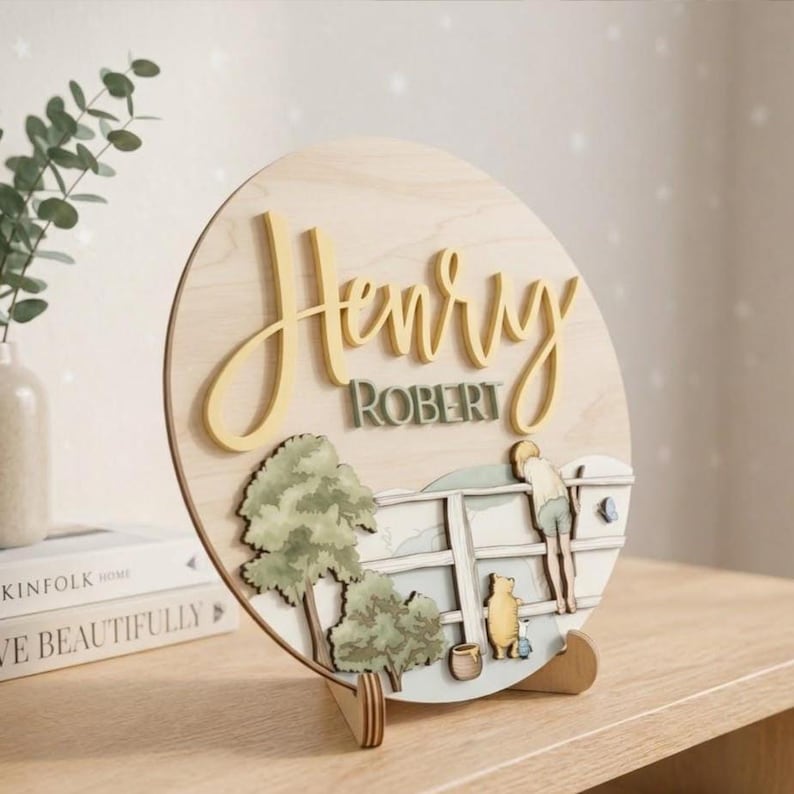 May include: A circular wooden sign featuring the name Henry Robert in raised yellow and green lettering. The design includes a layered wooden scene of a person and a bear leaning on a white fence near green trees, all set on a light wood base.