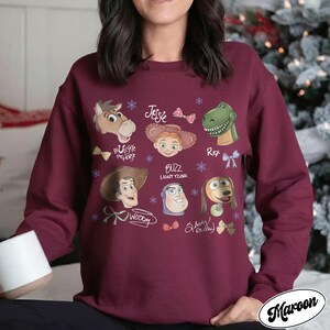 May include: Maroon sweatshirt featuring cartoon character illustrations including Woody, Buzz Lightyear, and Jessie. The design includes character names and decorative elements. The sweatshirt is a solid maroon color.