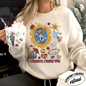 Puede incluir: Una sudadera de cuello redondo color crema con un estampado gr&aacute;fico de una persona con sombrero de vaquero y botas dentro de un marco dorado, rodeada de iconos como globos, una estrella con texto y la frase I KNEW IT, I KNEW YOU.