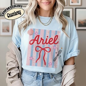 May include: Light blue t-shirt with the name "Ariel" in red script, a red bow, and a seashell graphic. The shirt has a striped design in pink and blue, with stars and a fork graphic. The shirt is labeled "Comfort Colors Chambray".