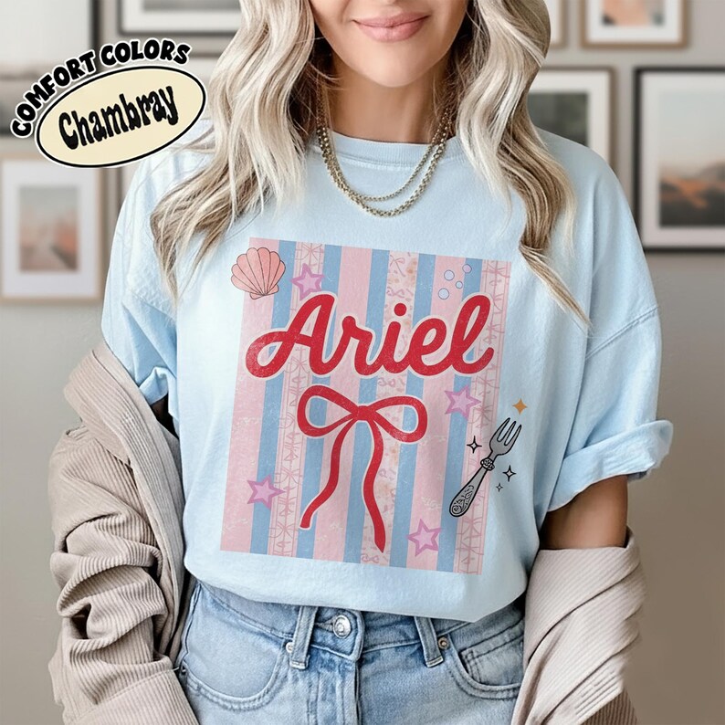 May include: Light blue t-shirt with the name "Ariel" in red script, a red bow, and a seashell graphic. The shirt has a striped design in pink and blue, with stars and a fork graphic. The shirt is labeled "Comfort Colors Chambray".