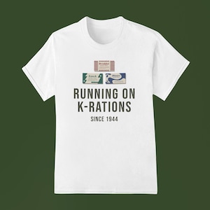 Puede incluir: Camiseta blanca con el texto negro "RUNNING ON K-RATIONS SINCE 1944" e ilustraciones de cajas de raciones K. Las cajas están etiquetadas como "Breakfast", "Lunch" y "Dinner".