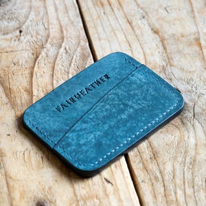 Op de afbeelding: Een teal leren kaarthouder met het woord "FAIRWEATHER" in reliëf op de voorkant. De kaarthouder heeft een getextureerde afwerking en witte stiksels rond de randen. De kaarthouder is rechthoekig en heeft een zwarte rand.