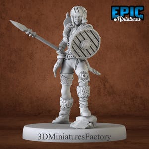 May include: A gray plastic miniature figurine of a female warrior holding a spear and a round shield. The figurine is standing on a gray base with the text "3DMiniaturesFactory" printed on it. The figurine is wearing a leather skirt, a fur-lined tunic, and leather boots. The shield has a raised design.
