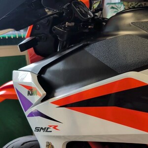 May include: Close-up of a motorcycle with a white, black, and orange color scheme. The motorcycle has the KTM logo and the letters SMC. The handlebars and mirrors are visible.