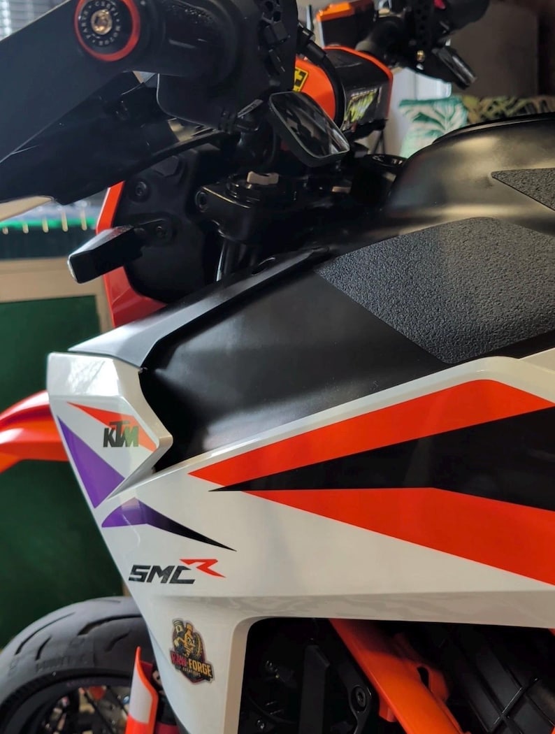 May include: Close-up of a motorcycle with a white, black, and orange color scheme. The motorcycle has the KTM logo and the letters SMC. The handlebars and mirrors are visible.