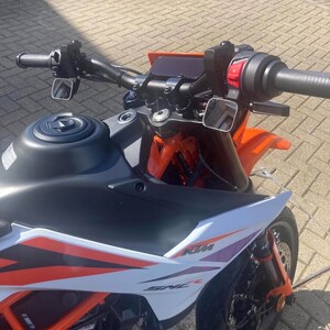 May include: A close-up view of a black and orange motorcycle. The motorcycle has a black handlebar, mirrors, and a fuel tank. The front fairing is white with orange and purple accents. The motorcycle's brand name, KTM, is visible.
