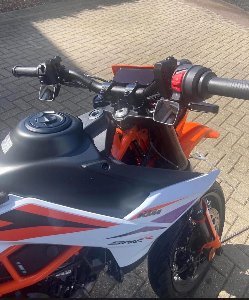 May include: A close-up view of a black and orange motorcycle. The motorcycle has a black handlebar, mirrors, and a fuel tank. The front fairing is white with orange and purple accents. The motorcycle's brand name, KTM, is visible.