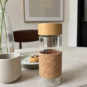 May include: A clear glass tea infuser bottle with a bamboo lid and a cork sleeve. The bottle has a stainless steel infuser basket inside. The bottle is sitting on a white tablecloth.